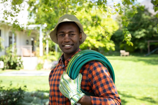 Retrato de um homem afro-americano no jardim usando um chapéu e luvas de jardinagem, carregando uma mangueira e sorrindo para a câmera. Família aproveitando o tempo em casa, conceito de estilo de vida