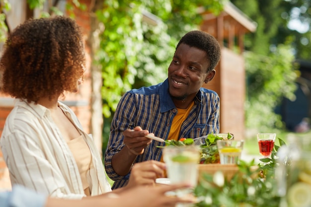 Retrato de um homem afro-americano sorridente, olhando para a namorada enquanto janta com amigos e família ao ar livre na festa de verão