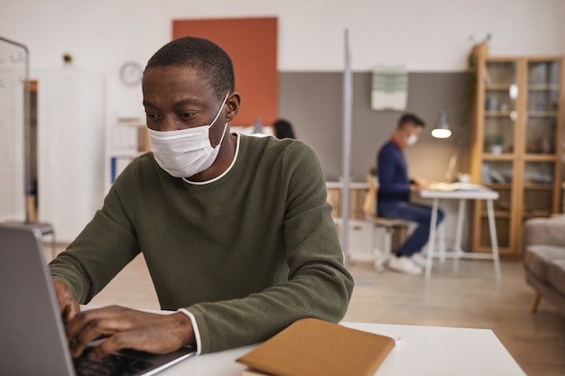 Retrato de um homem afro-americano usando máscara e usando laptop enquanto trabalha na mesa do escritório, copie o espaço