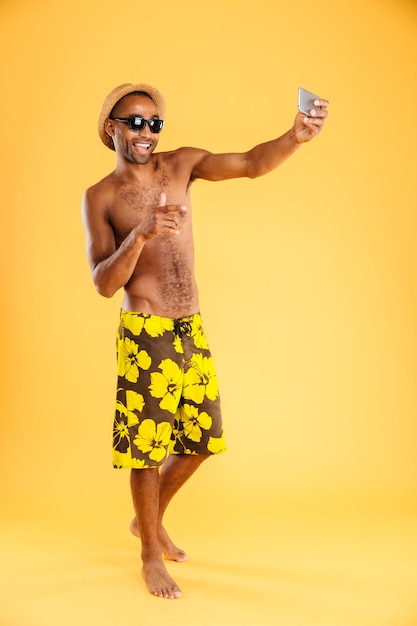 Retrato de um homem afro feliz em trajes de banho fazendo selfie foto em smartphone isolada em uma parede laranja