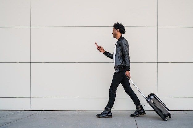 Retrato de um homem afro-turista usando seu telefone celular e carregando a mala enquanto caminha ao ar livre na rua
