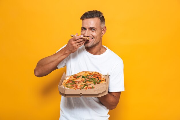 Retrato de um homem alegre de 30 anos em uma camiseta branca segurando e comendo pizza em pé isolado no amarelo