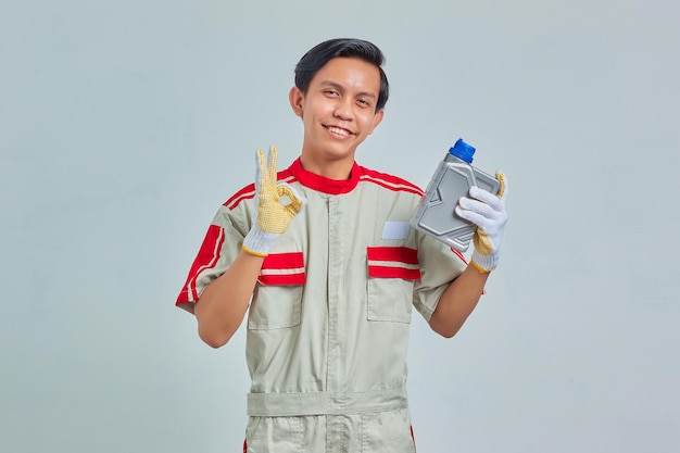 Retrato de um homem alegre e bonito vestindo uniforme de mecânico, segurando uma garrafa de plástico de óleo de motor e mostrando aprovação com o polegar para cima sobre um fundo cinza