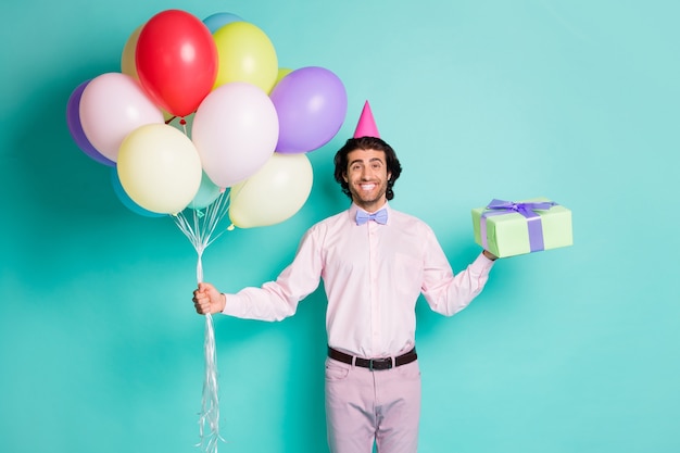 Retrato de um homem amigável segurando um gifbox de balões de hélio coloridos vestido em um cone de traje formal isolado em um fundo de cor turquesa