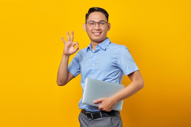 Retrato de um homem asiático bonito sorridente em óculos segurando laptop e gesticulando sinal de ok isolado no conceito de empresário e empresário de fundo amarelo