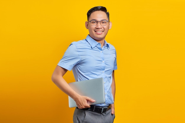 Retrato de um homem asiático bonito sorridente em óculos segurando laptop e olhando para câmera isolada no conceito de empresário e empresário de fundo amarelo