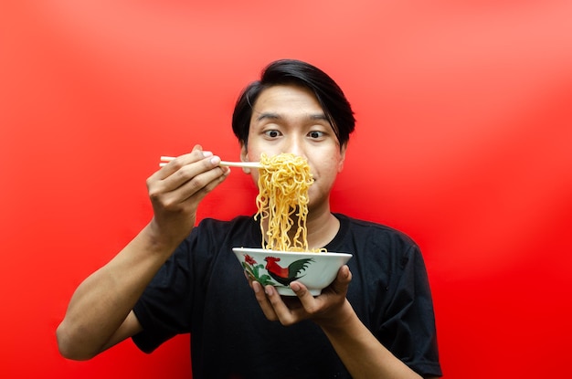 Retrato de um homem asiático feliz em camiseta preta come macarrão instantâneo usando pauzinhos e tigela com fome