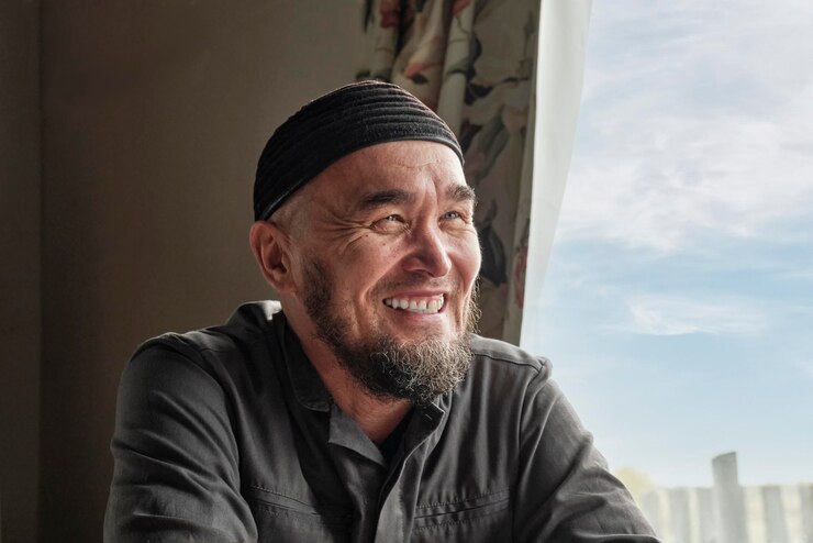 Foto retrato de um homem asiático idoso feliz com barba e boné sentado à janela e sorrindo