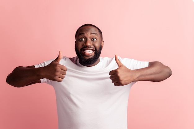 Retrato de um homem barbudo bonito afro-americano feliz mostrando o polegar para cima gesto na parede rosa