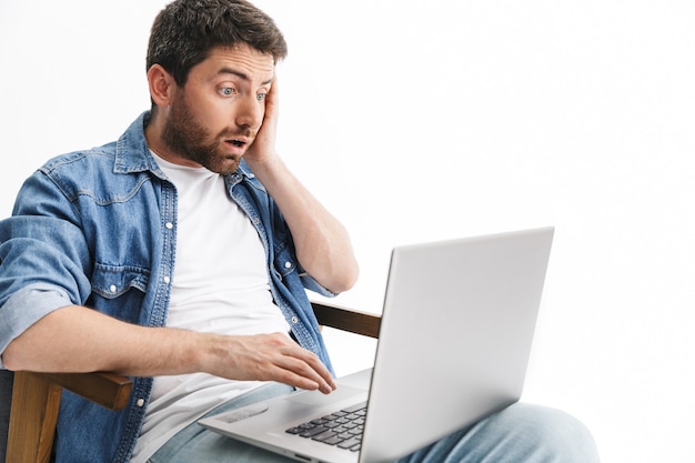 Retrato de um homem barbudo bonito chocado, vestindo roupas casuais, sentado em uma cadeira isolada na parede branca, trabalhando em um computador laptop