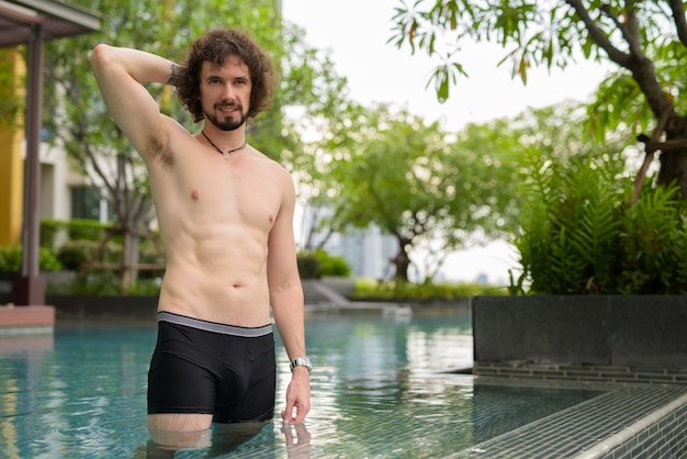 Retrato de um homem barbudo bonito com cabelo encaracolado sem camisa relaxando ao lado da piscina na cidade de Bangkok, Tailândia