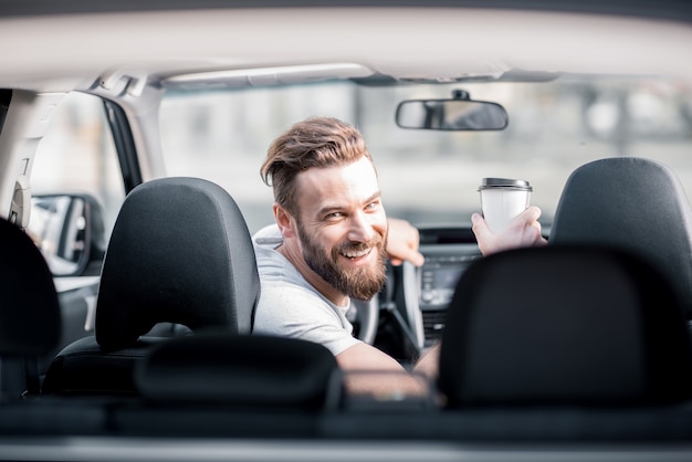 Retrato de um homem barbudo bonito olhando para trás, sentado no banco da frente do carro com uma xícara de café