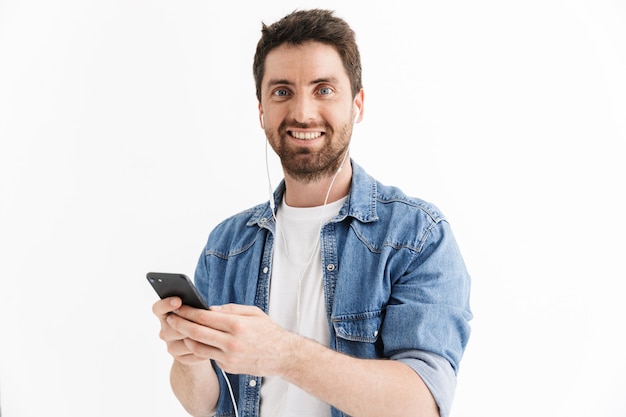 Retrato de um homem barbudo bonito vestindo roupas casuais, isolado, ouvindo música com fones de ouvido, segurando um telefone celular