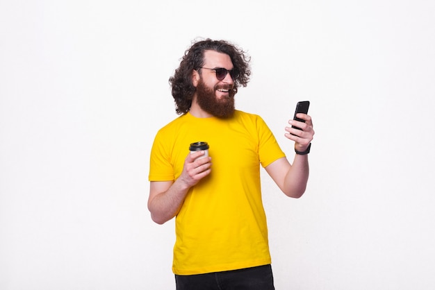 Retrato de um homem barbudo hippie com cabelo comprido encaracolado, bebendo uma xícara de café para ir e usando o smartphone