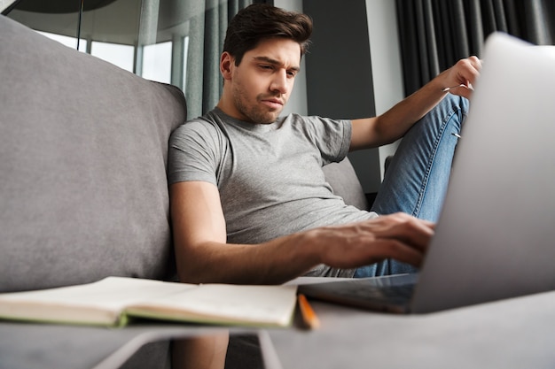 Retrato de um homem barbudo jovem sério e atraente, vestindo roupas casuais, sentado em um sofá na sala de estar, usando um laptop