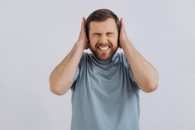 Retrato de um homem barbudo moderno de meia-idade em uma camiseta azul mostrando emoções cobrindo seus ouvidos em um fundo branco