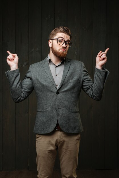 Retrato de um homem barbudo perplexo em óculos e terno apontando os dedos isolados no fundo preto de madeira