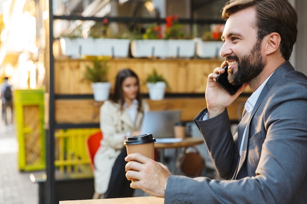 Retrato de um homem barbudo satisfeito vestindo jaqueta, bebendo café para viagem e falando no celular enquanto está sentado no café