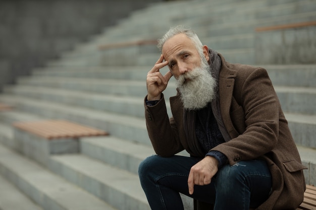 Retrato de um homem barbudo sênior ao ar livre, sentado em um banco em um parque.