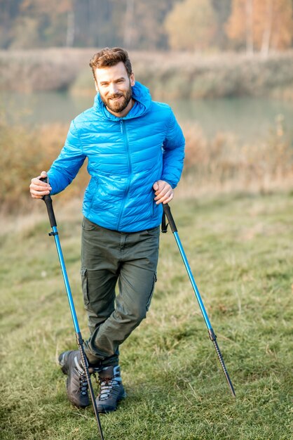 Retrato de um homem bonito com casaco azul, caminhando com varas ao ar livre perto do lago e da floresta