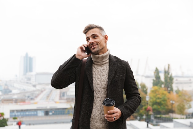 Retrato de um homem bonito dos anos 30 vestindo uma jaqueta, usando o celular, segurando um café para viagem, enquanto caminha pela rua da cidade