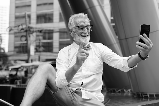 Retrato de um homem bonito e barbudo turista sênior vestindo roupas elegantes enquanto explora a cidade de Bangkok em preto e branco