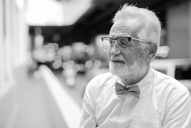 Retrato de um homem bonito e barbudo turista sênior vestindo roupas elegantes enquanto explora a cidade de Bangkok em preto e branco