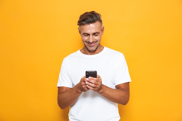 Retrato de um homem bonito e feliz posando isolado na parede amarela, usando telefone celular.