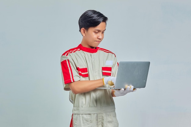 Retrato de um homem bonito e sorridente mecânico vestindo uniforme segurando e usando laptop em fundo cinza