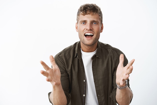 Retrato de um homem bonito feliz, animado e impressionado, gesticulando com as mãos sobre o corpo, sorrindo emocionado e intrigado ao contar sobre um produto incrível sobre a parede cinza