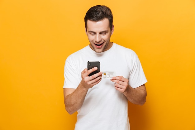 Retrato de um homem bonito feliz vestindo camiseta em pé, isolado na parede amarela, usando telefone celular, mostrando cartão de crédito