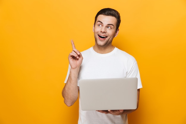 Retrato de um homem bonito feliz vestindo uma camiseta em pé, isolado na parede amarela, usando um computador laptop, comemorando, apontando para cima