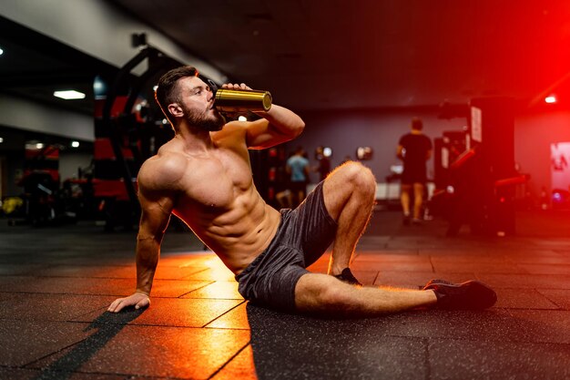 Retrato de um homem bonito fisiculturista seminu sentado no chão da academia Homem bebendo água