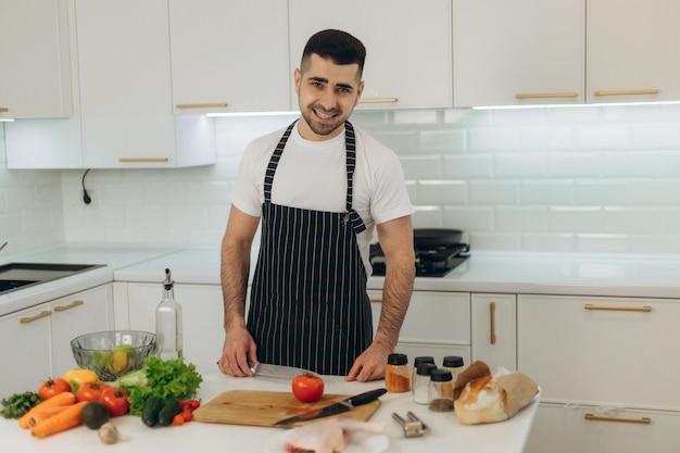 Retrato de um homem bonito na cozinha um homem vestido com um avental preto ele olha para a câmera na mesa da cozinha estão cozinhando produtos frango legumes especiarias