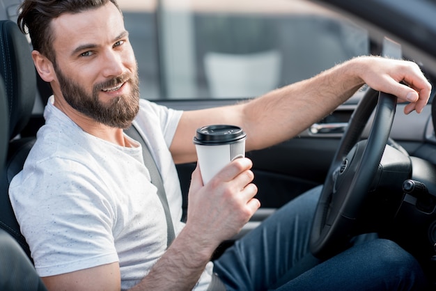 Retrato de um homem bonito vestido de cassual com camiseta branca dirigindo um carro com café para ir