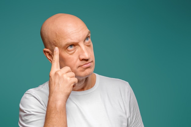 Retrato de um homem careca em uma camiseta branca na expressão pensativa, segurando um dedo perto de seu templo