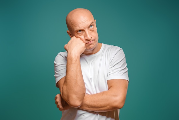 Retrato de um homem careca em uma camiseta branca na expressão pensativa, segurando um dedo perto de seu templo