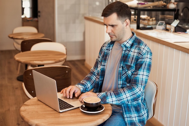 Retrato de um homem casual bonito de 35 anos sentado na mesa no café, trabalhando online com laptop