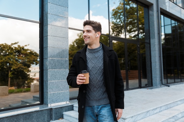 Retrato de um homem caucasiano dos anos 30 usando fones de ouvido segurando um copo de papel com café para viagem enquanto caminhava em frente a um prédio de vidro