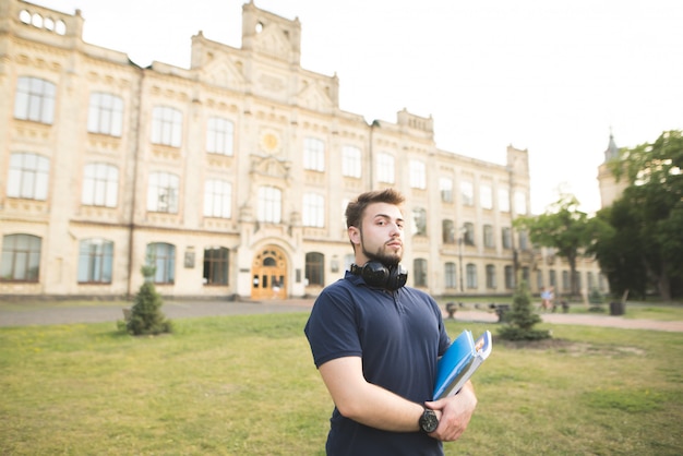Retrato de um homem com uma barba de pé em um campus com livros nas mãos e olhando para a câmera.