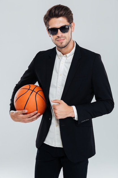 Retrato de um homem confiante e bonito com óculos escuros e terno preto segurando uma bola de basquete sobre a parede cinza