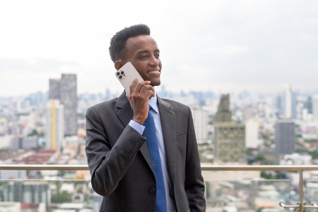 Retrato de um homem de negócios africano bonito de terno e gravata no telhado na cidade enquanto fala ao telefone