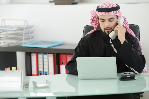 Retrato de um homem de negócios árabe inteligente usando laptop e falando ao telefone