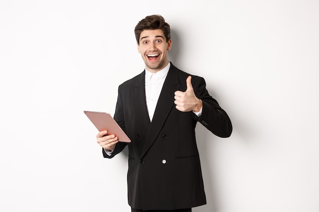 Retrato de um homem de negócios bonito de terno, mostrando o polegar para cima e fazendo um elogio, elogiando algo no tablet digital, em pé sobre um fundo branco