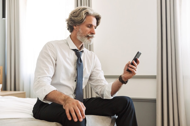 Retrato de um homem de negócios de cabelos grisalhos sênior concentrado sério, sentado na cama dentro de casa em casa usando o telefone celular.