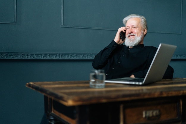 Retrato de um homem de negócios mais velho, de cabelos grisalhos, rindo, falando no celular, sentado à mesa com o computador portátil