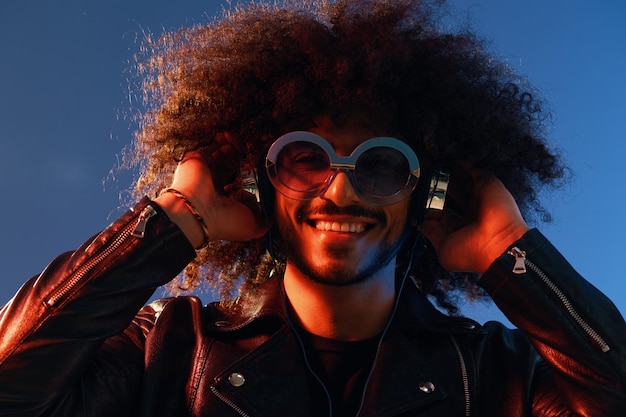 Retrato de um homem elegante com cabelos encaracolados com óculos e fones de ouvido em um fundo azul multinacional de cor clara casaco de couro preto tendência conceito moderno