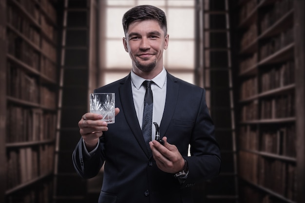 Retrato de um homem elegante de terno com um cachimbo de fumo e um copo na biblioteca. Conceito de negócios. Mídia mista