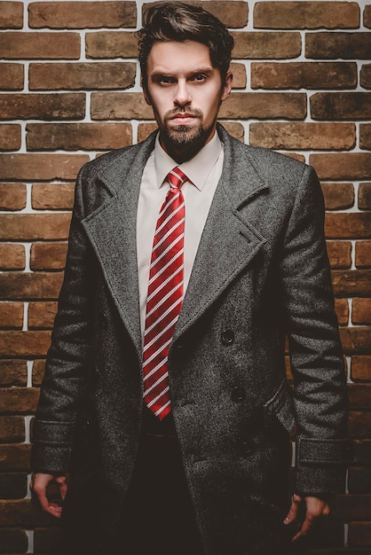 Foto retrato de um homem elegante e bem vestido, com barba, posando ao ar livre, olhando para longe, confiante e focado, homem maduro, de casaco, em pé sobre um fundo de parede de tijolos, modelo de moda elegante