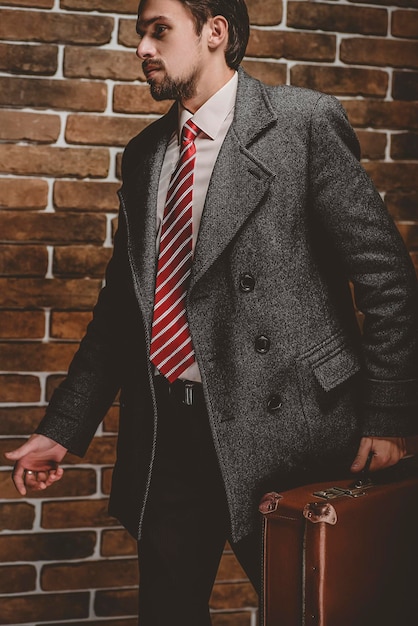 Foto retrato de um homem elegante e bem vestido, com barba, posando ao ar livre, olhando para longe, confiante e focado, homem maduro em pé de casaco com mala em um modelo de moda elegante de fundo de parede de tijolo